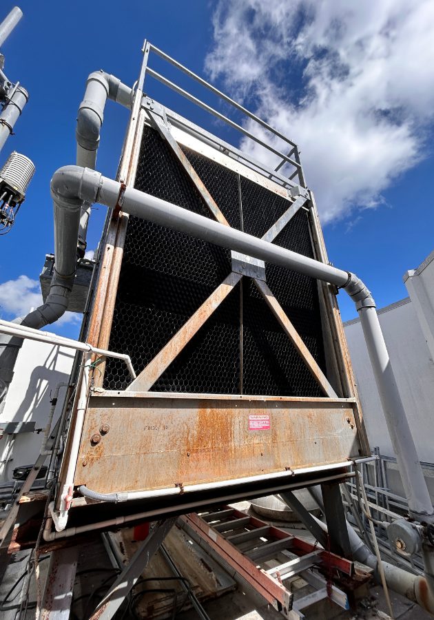 Industrial cooling/condenser unit with metal frame, rusted panels, and large gray pipes against a blue sky.