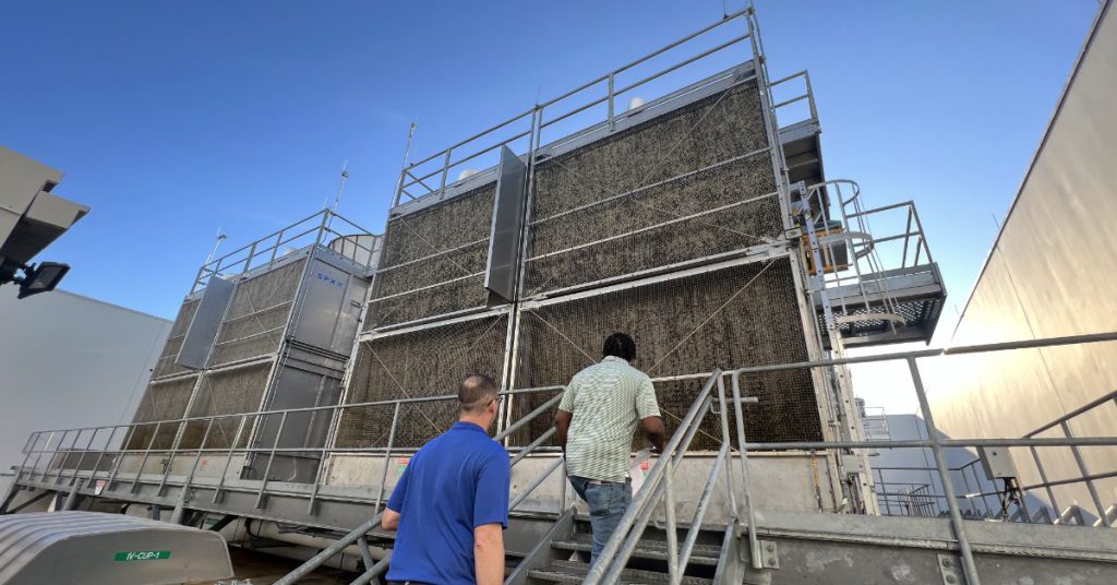 Two men walk up a metal staircase toward large industrial cooling towers on a rooftop.