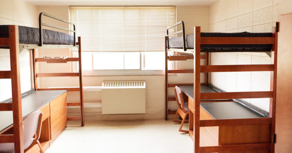 Dormitory room with two wooden bunk beds and desks, a window with blinds in the center.