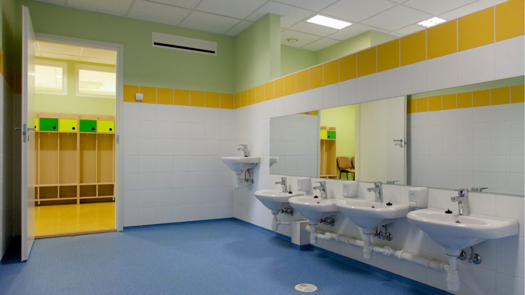 Public restroom sink area with five white basins, large mirror, yellow tile border, and blue floor; changing room with cubbies visible through open door.